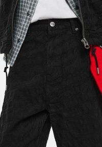 Pantaloni neri texture con vita alta, chiusura frontale con cerniera e bottone. Un dettaglio in tasca rossa aggiunge contrasto al design.