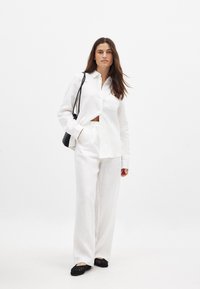 Balta lina pogu krekls ar kabatu, kombinēts ar brīviem baltiem lina biksēm un melniem austiem dzīvokļu apaviem. Modelis tur melnu somu.