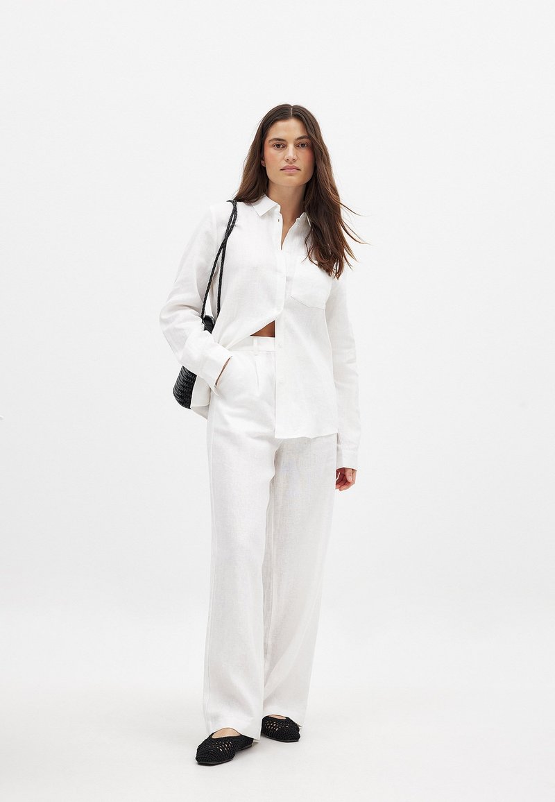 Balta lina pogu krekls ar kabatu, kombinēts ar brīviem baltiem lina biksēm un melniem austiem dzīvokļu apaviem. Modelis tur melnu somu.