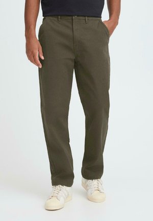 Homme portant un pantalon droit vert olive et des baskets blanches, debout avec les mains partiellement dans les poches.