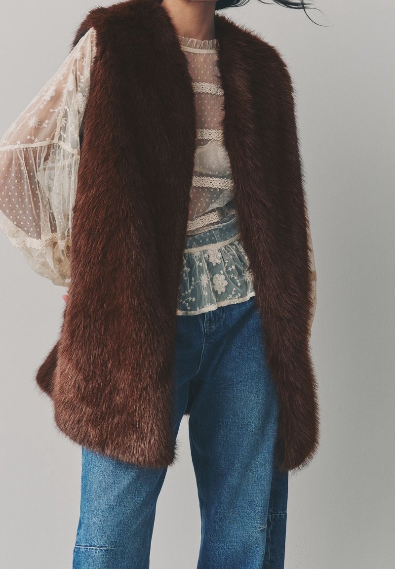 Femme portant un gilet en fourrure marron par-dessus une blouse blanche en dentelle transparente et un jean bleu, debout devant un fond gris uni.