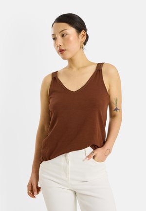 Mujer con cabello oscuro, con una camiseta marrón sin mangas y pantalones blancos, mostrando tatuajes en el brazo izquierdo y llevando pendientes y anillos de oro.