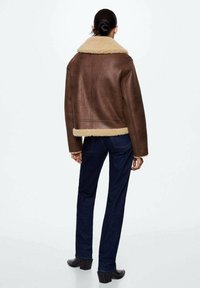 Brązowa kurtka ze shearlingu z beżowym futrzanym kołnierzem, skrócona konstrukcja i gładka tekstura. Połączona z ciemnoniebieskimi dżinsami i czarnymi botkami na obcasie.