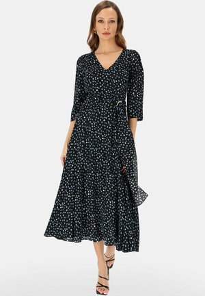 Kobieta ubrana w czarną midi sukienkę kopertową w niebieskie kropki, z rękawami do łokcia i czarne sandały na wysokim obcasie z paskami, stojąca na jednolitym tle.