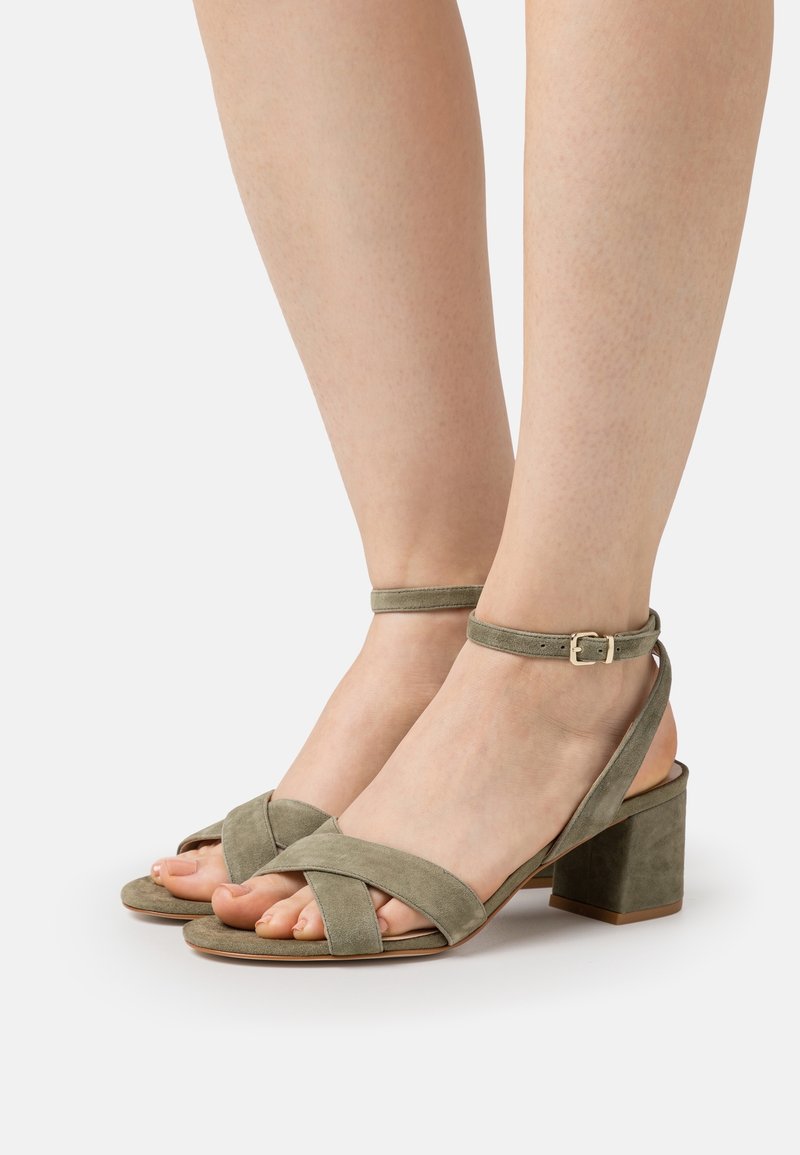 Pieds portant des sandales à talons blocs en daim vert olive avec des brides à la cheville et des brides entrecroisées sur les orteils, sur fond blanc.