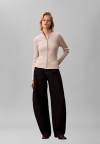 Jersey beige acanalado con cremallera combinado con vaqueros negros de pierna ancha, que presenta una textura suave y mangas largas. Botines marrones completan el conjunto.