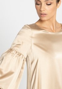 Sukienka satynowa w delikatnym beżowym odcieniu z okrągłym dekoltem i długimi, puszystymi rękawami z marszczonymi mankietami. Gładka tekstura i elegancki układ.