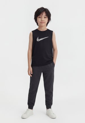Chłopiec stoi zwrócony przodem, ubrany w czarną koszulkę bez rękawów marki Nike, ciemnoszare spodnie dresowe i białe trampki na białym tle.