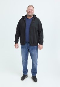 Uomo di mezza età in piedi, indossa una felpa nera con zip, una maglietta blu, jeans blu e sneakers nere, sorridente su uno sfondo chiaro e uniforme.