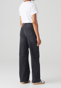 Vaqueros de tiro alto, de pernera ancha y de lavado oscuro, confeccionados en denim, con dos bolsillos traseros. Combinados con una camiseta blanca básica y zapatos informales.