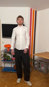 Jeune homme en chemise blanche et pantalon noir debout dans une pièce avec un mur à rayures, une télévision, une lampe orange, une bibliothèque, une plante et une valise décorée d'autocollants.