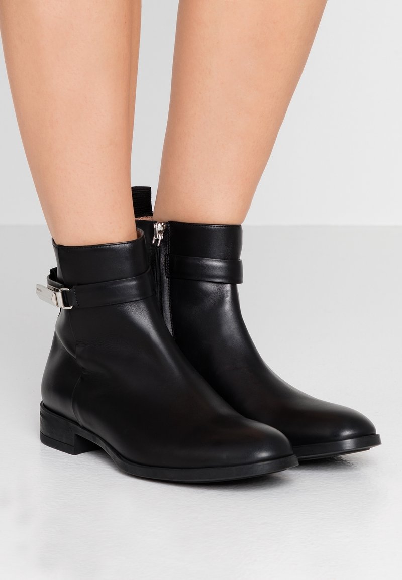 Bottines en cuir noir avec fermeture éclair latérale, sangle et boucle argentée portées sur des jambes nues sur un fond neutre.