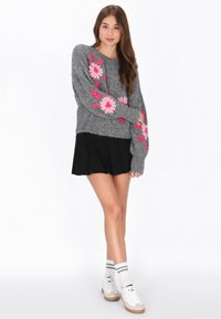 Grå stickad tröja med rosa blommig broderi på ärmarna, kombinerad med en svart plisserad kjol, vita strumpor och avslappnade sneakers.