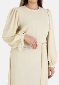 Cream-colored dress with long, puffed sleeves and wrist ties. Smooth texture and a simple round neckline, cinched at the waist with a belt.