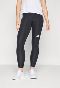 Schwarze Leggings mit einem weißen Drei-Streifen-Logo, enganliegendem Design, glatter Textur und Seitentaschen. Tragen mit einem hellen Oberteil.