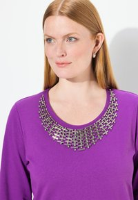 Lila Langarmshirt mit einem dekorativen Design aus silbernen Perlen und Stäben am Ausschnitt. Glatte Textur, runder Ausschnitt und taillierte Passform.