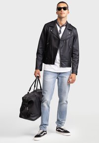 Svart lær motorsykkeljakke med sølvglidelåser, hvit t-skjorte, lyseblå jeans, svart duffelbag og svarte sneakers med hvite såler.