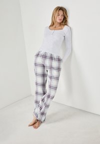 Grijze henley met lange mouwen, gecombineerd met geruite pyjamabroek in paars, blauw en wit. De broek heeft een elastische tailleband en zijzakken.