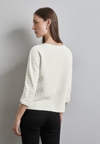 Weißer gesteppter Pullover mit einer kastigen Form, langen Ärmeln, die an den Bündchen aufgerollt sind, und einem runden Ausschnitt. Getragen mit schwarzen Slim-Fit-Jeans.