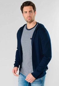 Marineblauwe rits-hoodie met een soepele textuur, lange mouwen en capuchon, gedragen over een lichtgrijze gestreepte T-shirt. Casual spijkerbroek.