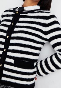 Svartvit randig kofta gjord av mjukt, strukturerat tyg. Har knappstängning, rund halsringning och en framficka.