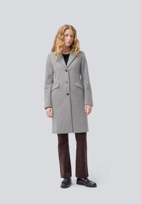 Palton gri din lână cu un design croit, având rever cu tăietură, două buzunare frontale și nasturi negri. Purtați peste pantaloni și pantofi maro închis.