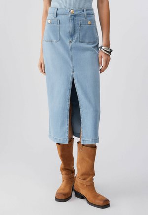 Gonna midi in denim azzurro chiaro con spacco frontale, due tasche applicate frontali con bottoni dorati, abbinata a stivali in camoscio marrone a metà polpaccio e braccialetti argento.