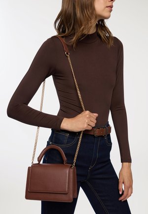 Borsa in pelle marrone con catena dorata e manico superiore. Texture liscia, forma rettangolare e dettaglio con pattina. Indossata con jeans scuri.