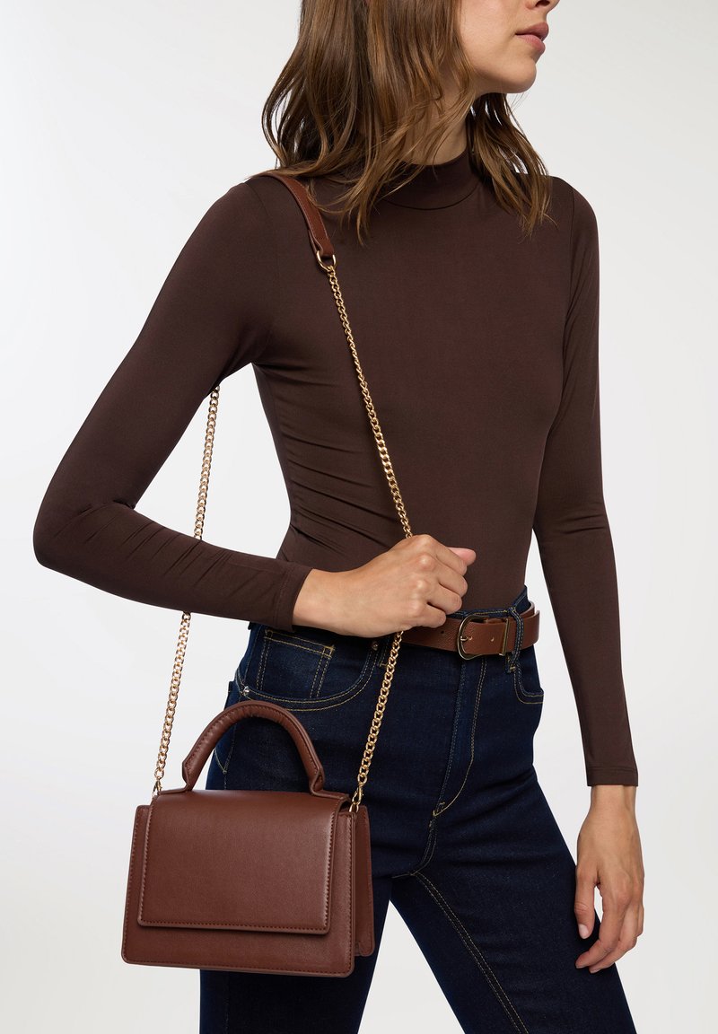 Borsa in pelle marrone con catena dorata e manico superiore. Texture liscia, forma rettangolare e dettaglio con pattina. Indossata con jeans scuri.
