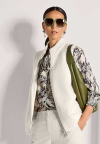 Frau mit übergroßen grünen Sonnenbrillen, einem Schlangenhautmuster-Bluse, einem weißen strukturierten Westen, hellen Hosen und einer olivgrünen Leder-Schultertasche.