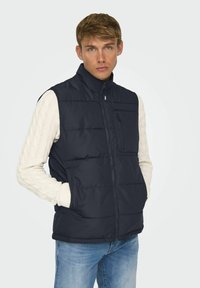 Gilet matelassé bleu marine avec un col haut, une fermeture éclair à l'avant et une poche poitrine ; présente un motif matelassé et un tissu uni ; superposé sur un pull en laine torsadée crème.