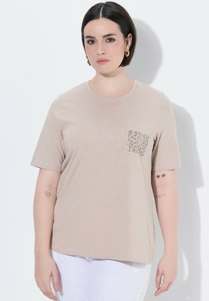Kobieta z krótkimi ciemnymi włosami, ubrana w beżowy t-shirt z kieszenią w panterkę i białe spodnie, stojąca na białym tle.