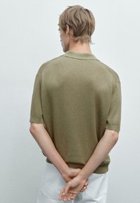 Polo corto a maniche corte di colore khaki, con un motivo testurizzato, colletto tondo e dettagli a coste, visto da dietro con le mani incrociate.