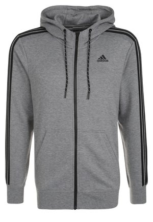 Mężczyzna poprawiający szarą bluzę Adidas zapinaną na zamek, ubrany w pasujące szare spodnie dresowe, niebieską koszulkę Adidas i szare sneakersy z pomarańczowymi akcentami.