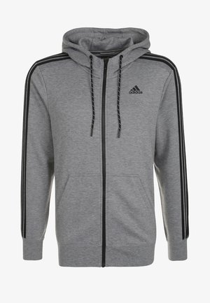Homme ajustant un sweat à capuche zippé gris Adidas, portant un pantalon de survêtement gris assorti, un t-shirt bleu Adidas et des baskets grises avec des touches orange.
