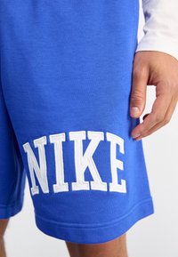 Niebieskie bawełniane szorty z białym haftowanym logo "NIKE" po lewej stronie, o gładkiej fakturze i prążkowanym dole. Luźny sportowy styl.