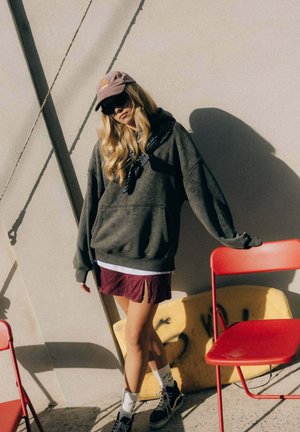 Młoda kobieta ubrana w za dużą ciemną bluzę z kapturem, bordową spódnicę, trampki, czapkę i okulary przeciwsłoneczne, oparta o czerwone składane krzesło przy słonecznej ścianie.