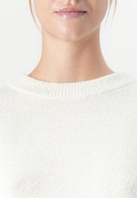 Parte inferiore del volto e collo di una persona che indossa un maglione a maglia bianco, con labbra rosa lucide visibili su uno sfondo chiaro.