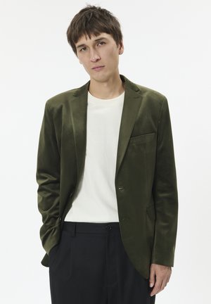 Olivgrüner Samt-Blazer mit Revers, einreihiger Knopfverschluss und Seitentaschen, getragen über einem weißen Hemd und kombiniert mit schwarzen Hosen.