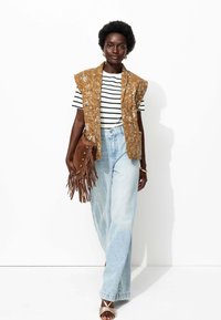 Femme avec les cheveux courts et bouclés portant une chemise rayée, un gilet floral moutarde, un jean bleu clair, tenant un sac à franges marron et des sandales dorées.