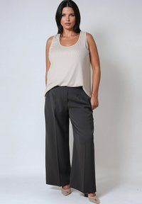 Femme aux cheveux noirs portant un haut sans manches beige, un pantalon noir à jambes larges et des escarpins beiges à bout pointu, se tenant contre un fond clair et uni.