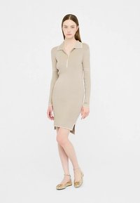 Beżowa sukienka z prążkowanego dzianiny z suwakiem przy kołnierzu, długimi rękawami, bocznymi rozcięciami i białymi ozdobnymi lamówkami. Modelka ma na sobie złote płaskie buty.