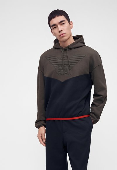 Jeune homme portant un sweat à capuche bicolore marron et marine avec le logo GA, debout, une main dans la poche, devant un fond clair uni.