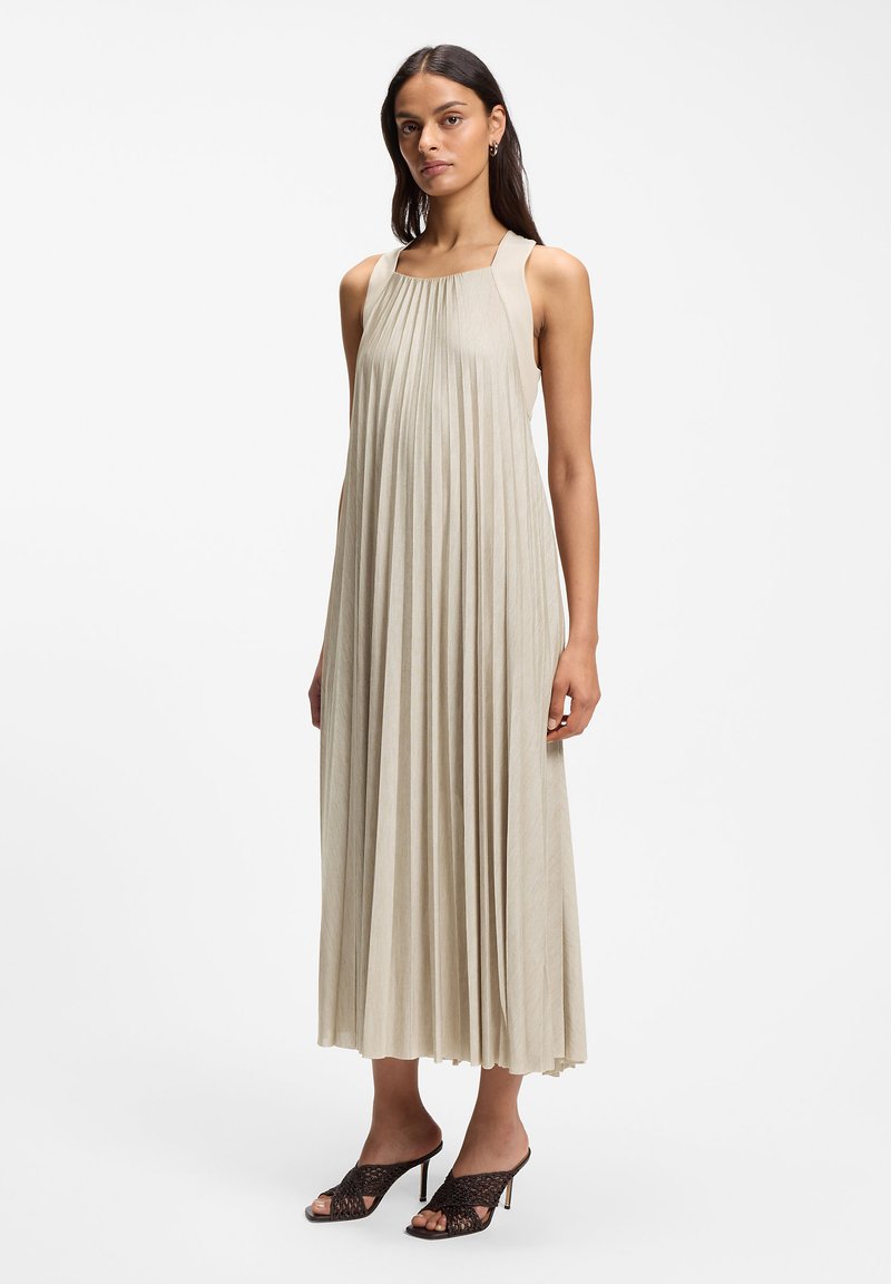 Femme portant une robe midi plissée beige sans manches et des sandales noires à talons ouvertes, se tenant devant un fond blanc uni.