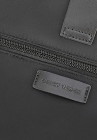 Sort stof håndtaske med en glat lynlås lukning og et lærred prægede mærke med teksten "GERRY WEBER." Tekstureret overflade med solid syning.