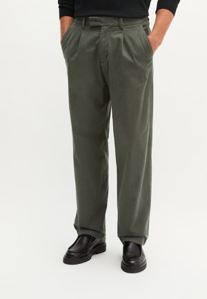 Homme portant un pantalon plissé vert olive avec des chaussures noires et les mains dans les poches, sur un fond clair uni.