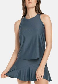 Vrouw in een mouwloos blauwe sporttop en bijpassende flared tennisrok, die de zoom van de rok met één hand vasthoudt.