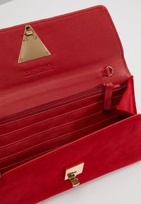 Punainen mokkanahkainen lompakko, jossa kultainen kolmion muotoinen koriste, sisällä vetoketjutasku, useita korttipaikkoja ja napsautettava suljin. Merkattu VALENTINO-logolla.