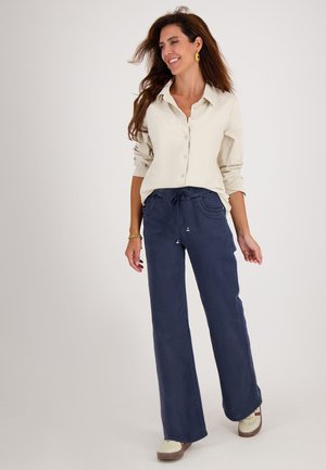 Beige overhemd met opgerolde mouwen, gecombineerd met marineblauwe wijde broek met een trekkoord in de taille en zijzakken. Lichte schoenen.