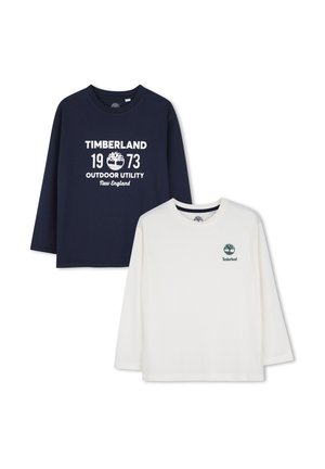 Twee Timberland longsleeve shirts, één marineblauw met wit logo en tekst, één wit met klein groen logo op de borst, plat weergegeven.